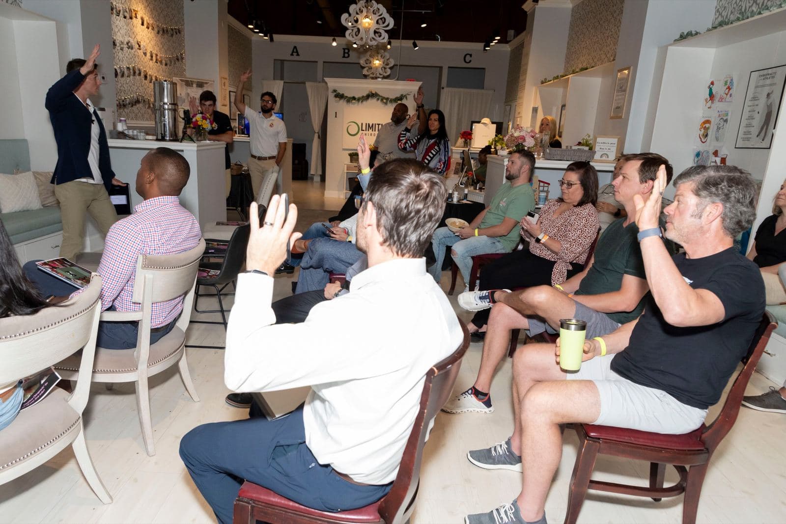 About a dozen guests seated in the Limitless Chiropractic lobby with hands raised during an orientation
